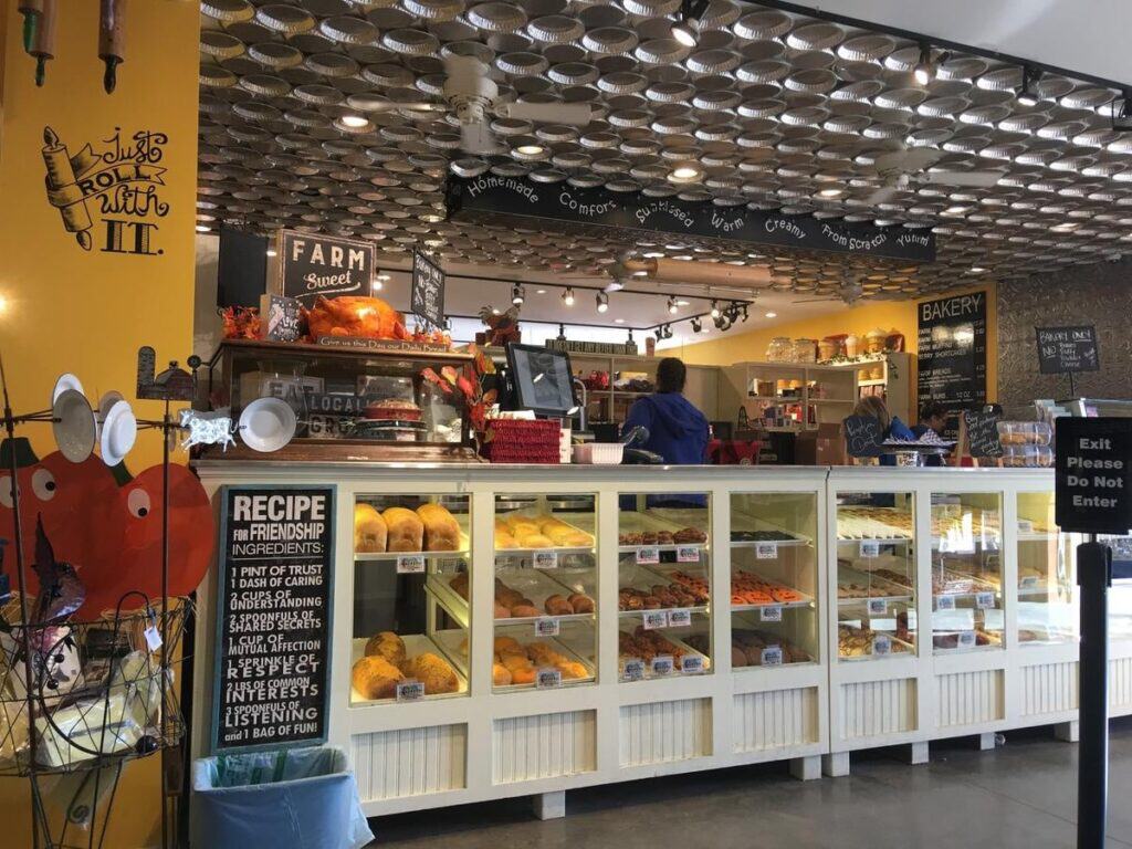 Vintage white display cases brim with fresh loaves, pies, and cookies, flanked by a chalkboard “Recipe for Friendship” and other hand-lettered signs. Pie tins tiled across the ceiling and farm-style décor create a cozy, from-scratch atmosphere that invites customers to eat local.