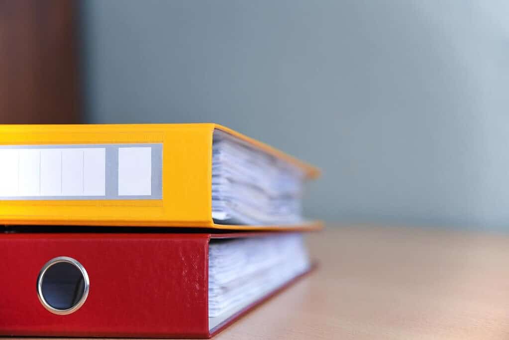 Two thick binders, one red and one yellow, stacked on a wooden surface, each stuffed with neatly filed papers and labeled spines. Keeping separate binders for topics like herbal remedies and emergency prep helps streamline information and reduce overwhelm on the homestead.